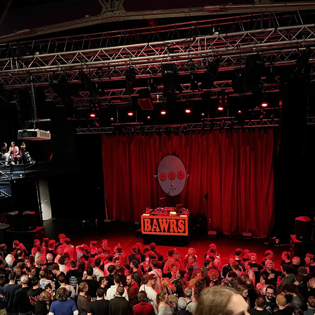 Aufnahme einer Konzertbühne mit DJ-Tisch und einem „BAWRS“-Schild sowie einem dreiäugigen blauen Kopf als Logo im Hintergrund.