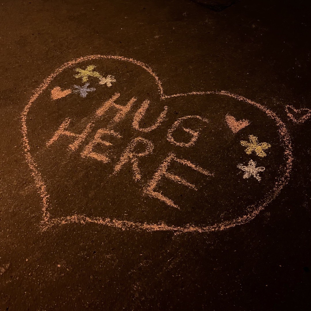 Ein mit Kreide gemaltes Herz auf einer Straße. Darin steht mit Blumen verzieht "Hug here".
