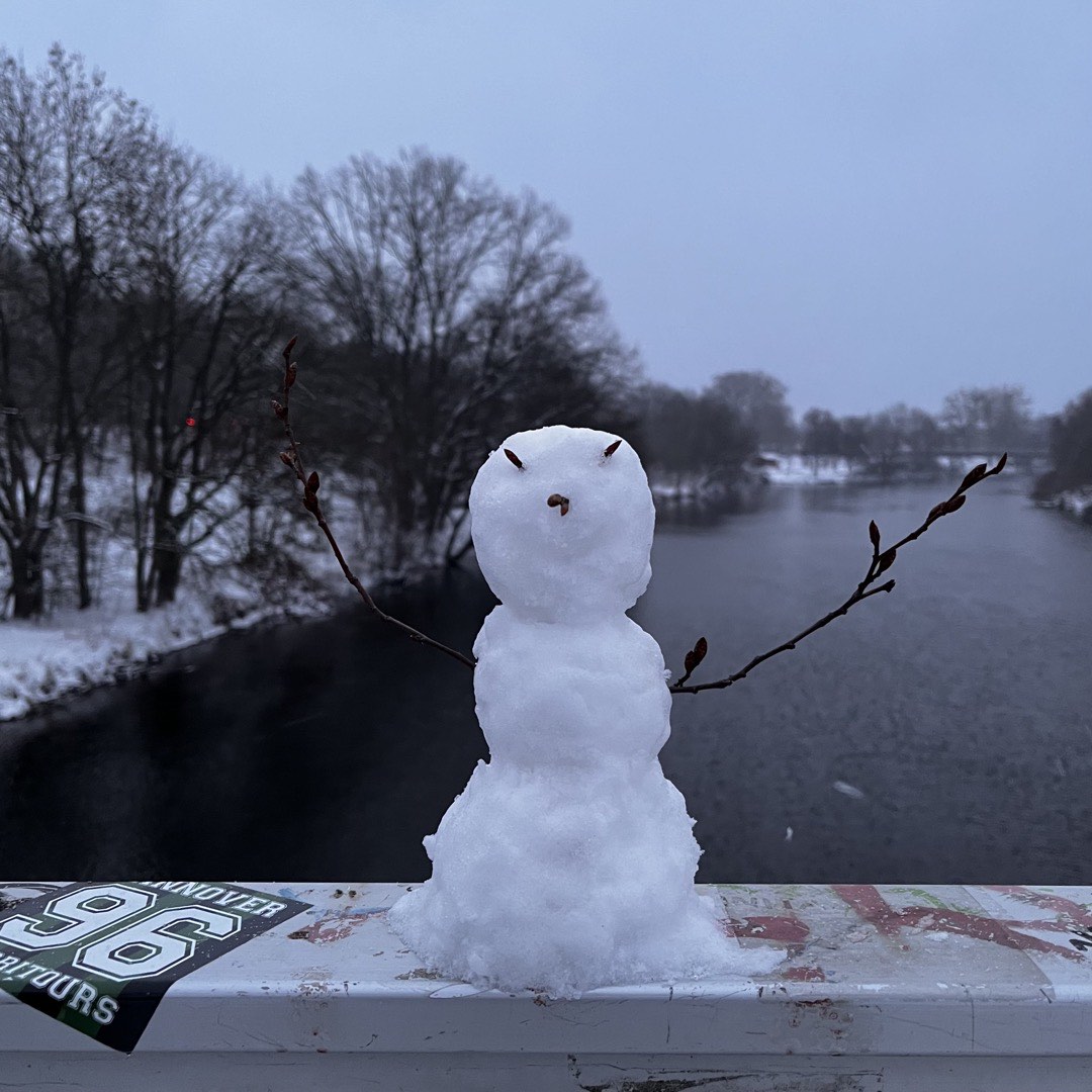 Eine sehr kleine Schneeperson auf einem Geländer auf einer Brücke an einem Fluss.