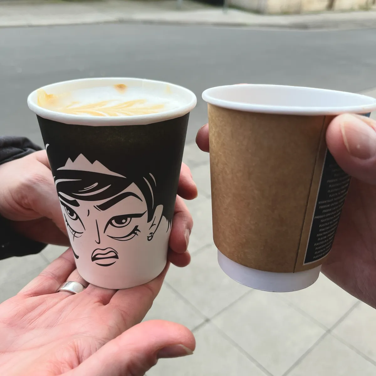 Zwei Kaffeebecher to go. Der linke Becher ist mit einer abstrakten Zeichnung eines grimmigen Gesichts verziert.
